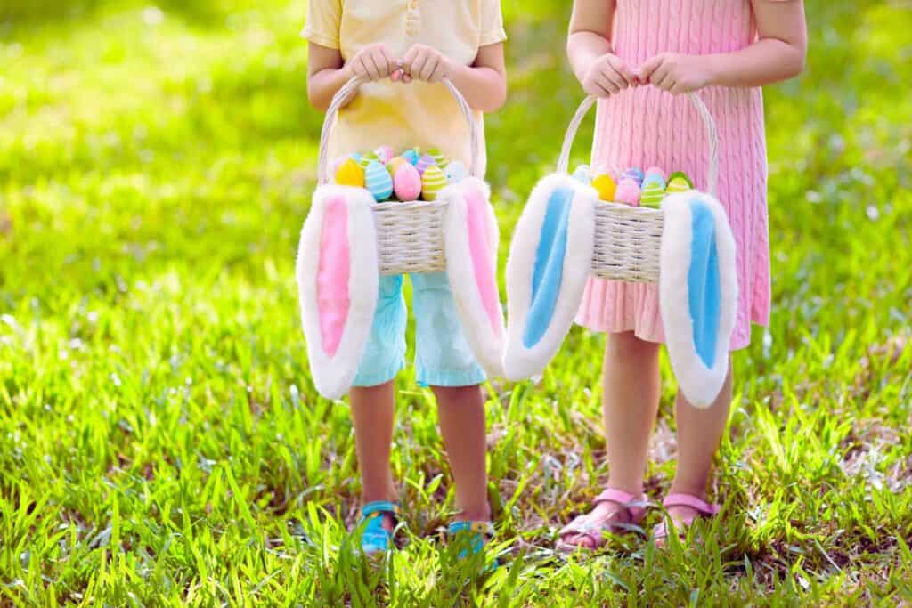 2 kids in a sunny Spring garden holding Easter baskets filled with Easter eggs and floppy bunny ears hanging out