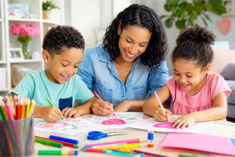A mother and her children coloring Mother's Day pages together at a table surrounded by art supplies.