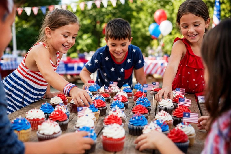 Children reaching for red, white, and blue mini cupcakes arranged on a table at an outdoor 4th of July party.