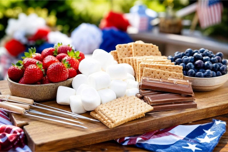 A wooden board with strawberries, blueberries, marshmallows, graham crackers, and chocolate arranged for making s'mores.