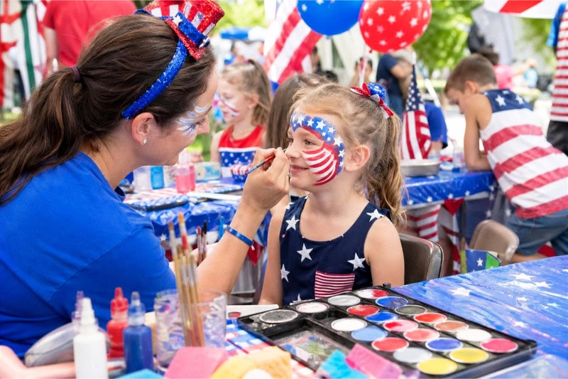 A child having their face painted with stars and stripes designs at a festive 4th of July party face paint station.