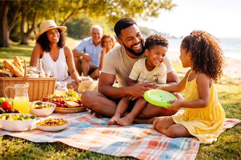 A family enjoying a sunny outdoor picnic with a father and children playing and sharing food at a park or beach.
