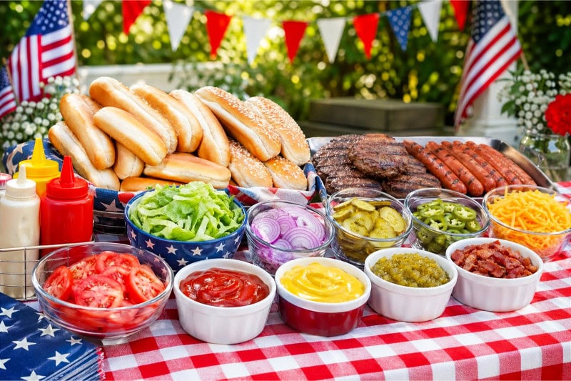 A picnic table with bowls of hot dog and burger toppings including lettuce, tomatoes, pickles, onions, cheese, and condiments, along with buns, hot dogs, and burgers, decorated with small American flags.