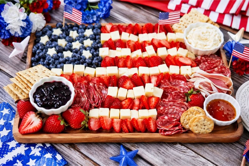 A charcuterie board with strawberries, blueberries, cheese, cured meats, and crackers arranged in patriotic colors on a wooden table.