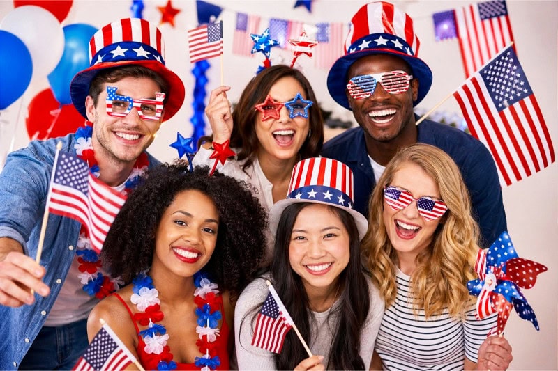 A group of people posing with American flag props at a 4th of July photo booth decorated with red, white, and blue party decorations.