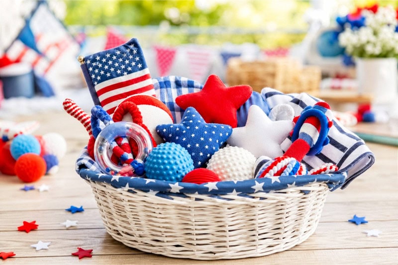 A basket filled with red, white, and blue sensory toys for babies and toddlers placed on a wooden surface with small American flags and star decorations around it.