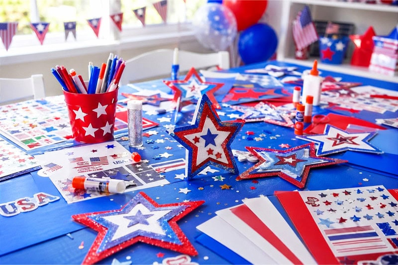 A craft station with red, white, and blue construction paper, patriotic stickers, and craft supplies arranged on a table for a kids' 4th of July party.
