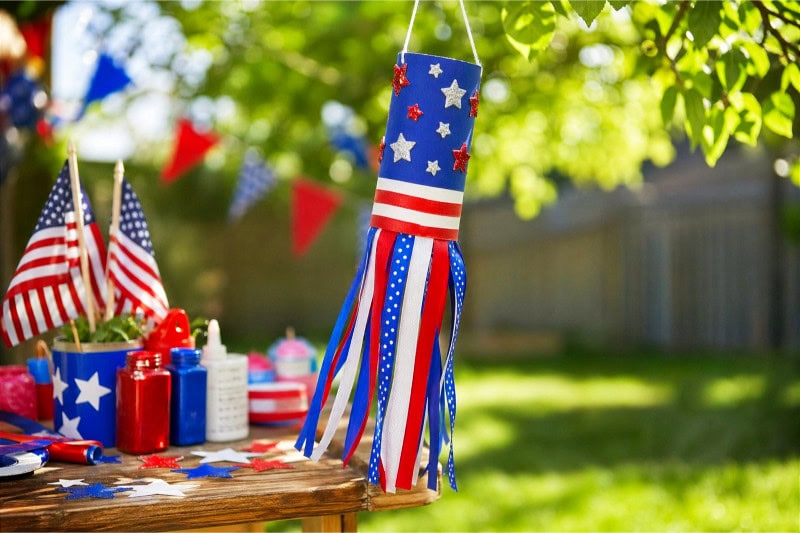 A colorful 4th of July windsock craft made from a toilet paper roll hanging outside with patriotic decorations around it.
