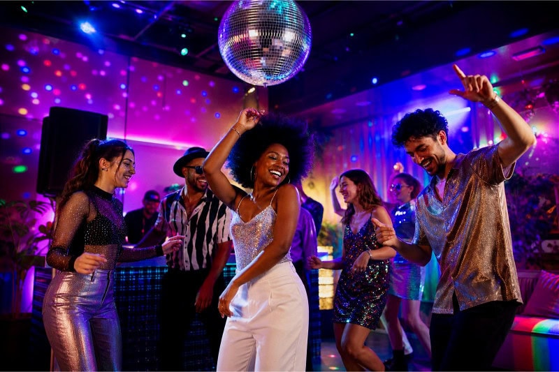 People dancing and enjoying a colorful indoor disco party with a disco ball and vibrant lights.