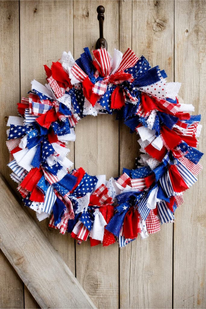 A colorful scrap fabric rag wreath with red, white, and blue fabrics hanging on a wooden surface.