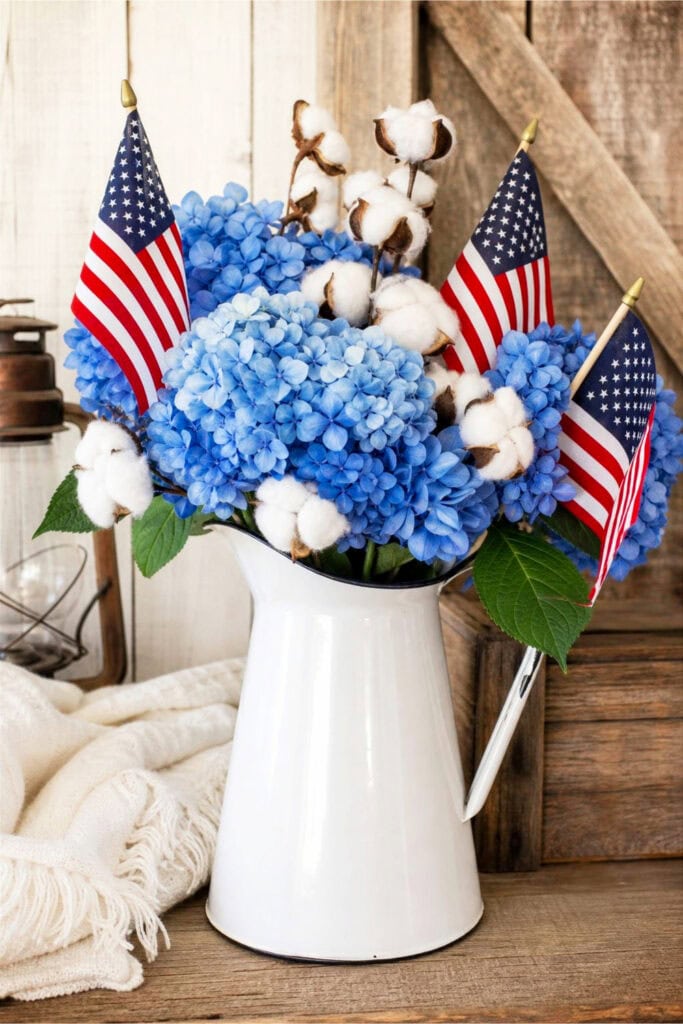White enamel pitcher holding blue hydrangeas, cotton stems, and small American flags on a wooden surface.