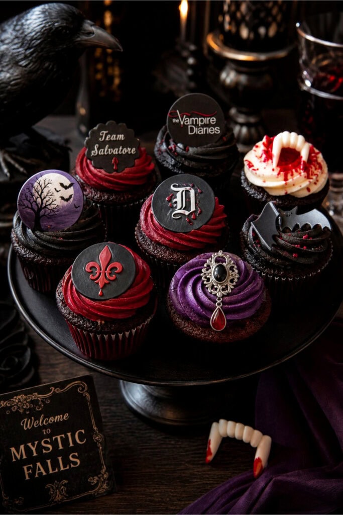 A display of cupcakes decorated with vampire-themed toppers, including a crow figurine and vampire fang decoration on a dark wooden table.