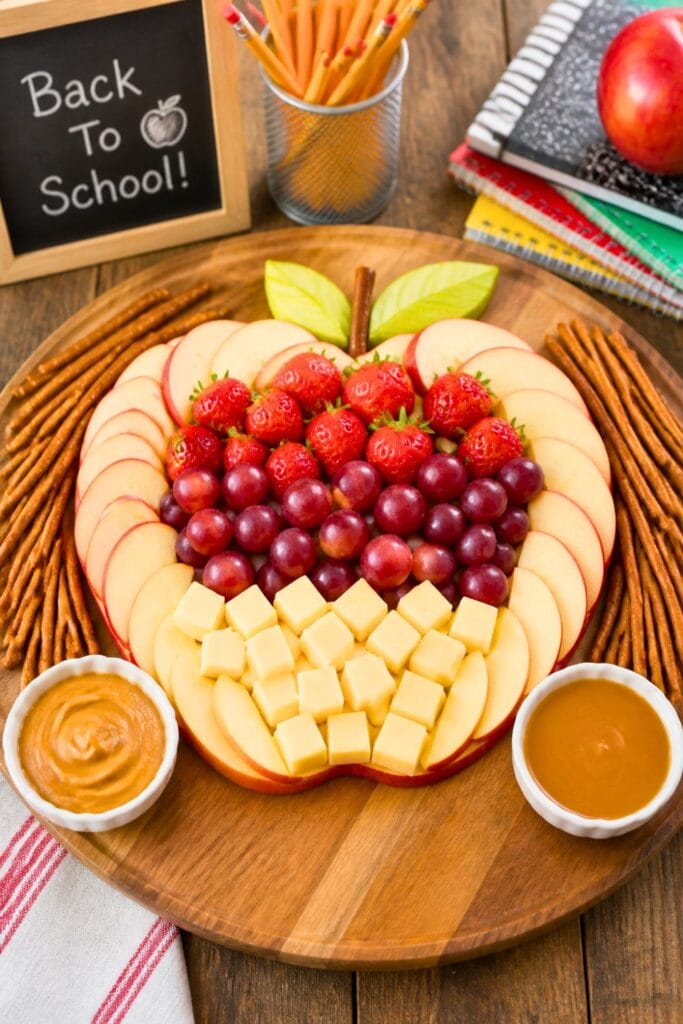 A snack board with apple slices, grapes, pretzel sticks, and dips on a wooden table with school supplies in the background.