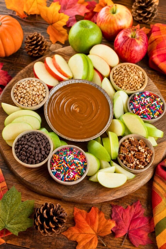 A wooden board with sliced apples, caramel dip, and various toppings arranged for a fall snack.