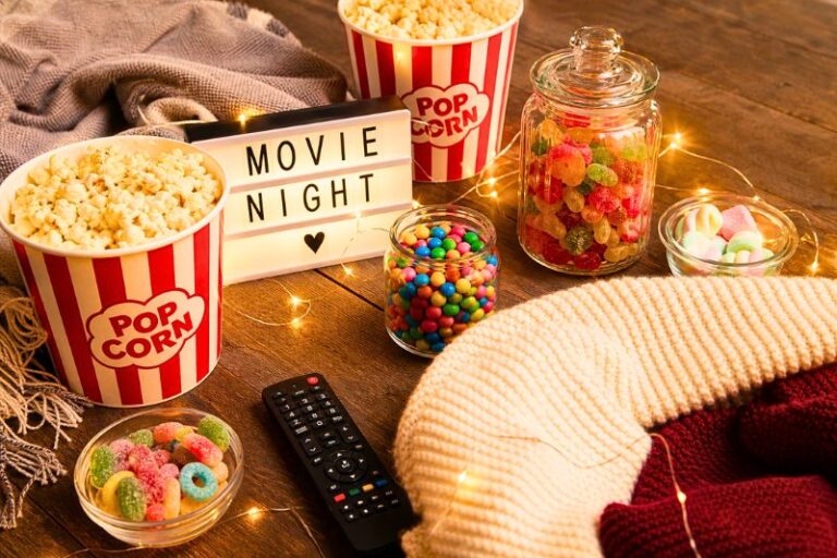 A styled flat-lay turned slight angle of a movie night setup: popcorn buckets, candy jars, remote, cozy blankets, and fairy lights woven through. Bright, saturated colors with intentional spacing so each item pops. Background neutral wood or soft fabric.