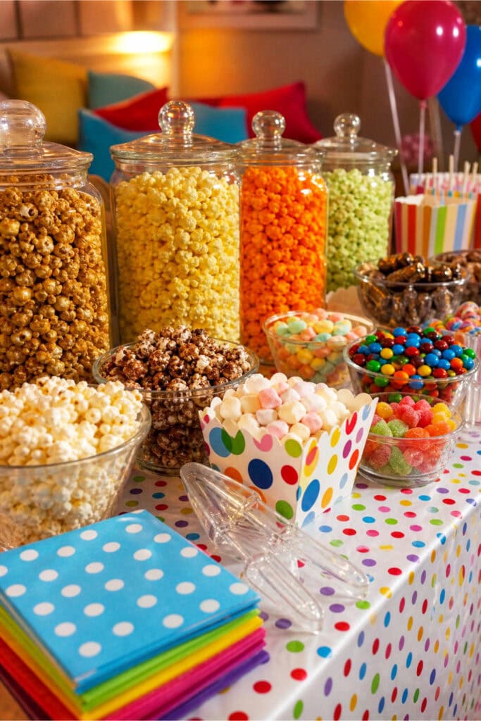 A popcorn bar with different popcorn flavors, colorful candies, and mini marshmallows arranged on a decorated table for a kids' movie night party.