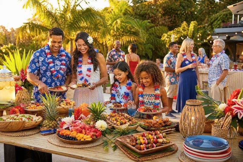 A diverse group of people enjoys a festive tropical Labor Day luau outdoors with leis, fruit, flowers, and patriotic decorations.
