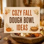 A rustic table with a styled autumn-decorated wooden dough bowl filled with pumpkins, gourds, pinecones, fairy lights, and seasonal accents.